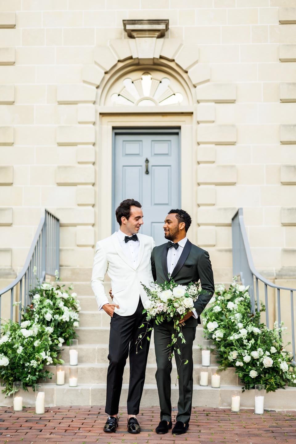 interracial couple two grooms wearing tuxedo one is holding a bouquet of white flowers and greenery standing in front of stone staircase decorated with matching florals and candles